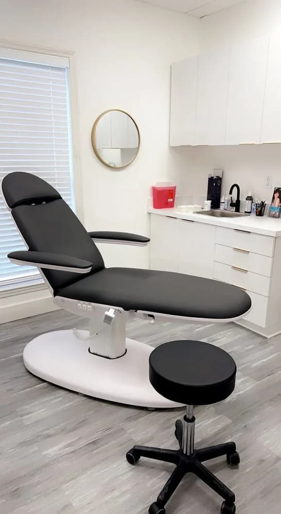 Treatment room with black chair, round gold-framed mirror, clean white cabinetry